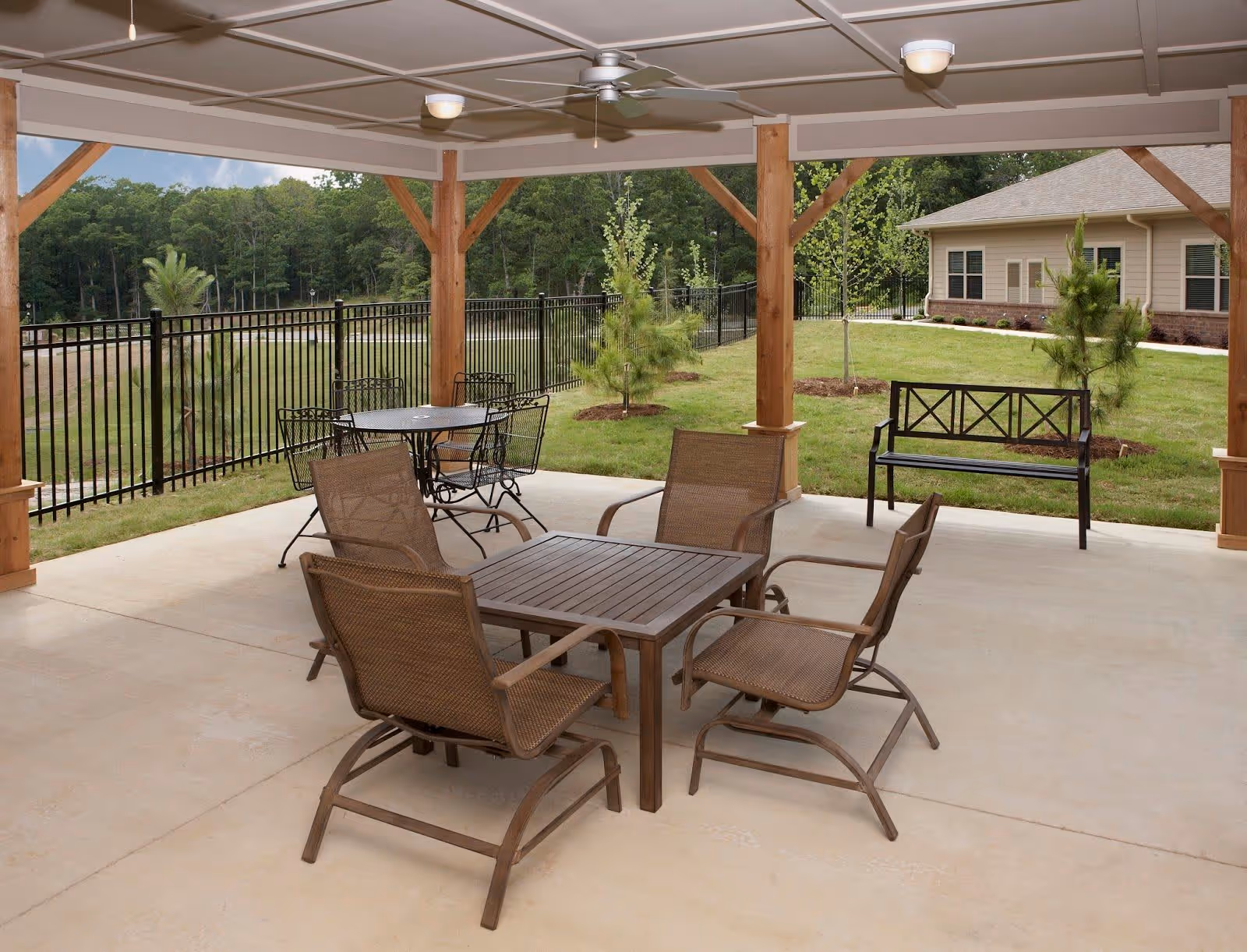 Covered outdoor patio area with a wooden table and four mesh chairs, a round metal table with four matching chairs, a black metal bench, ceiling fans, and a view of a grassy yard with small trees and a building in the background.