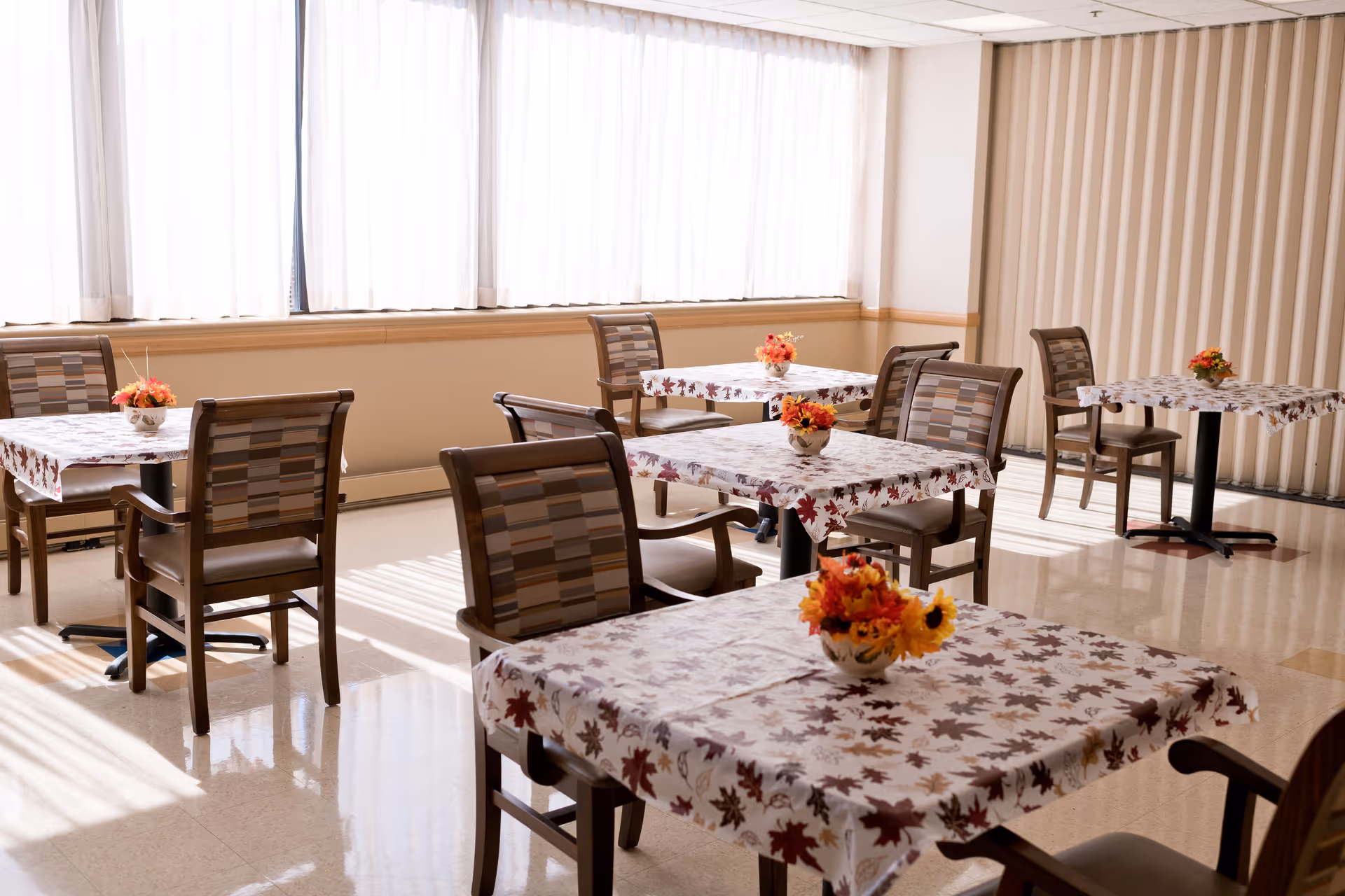 A bright dining room with several tables covered in autumn-themed tablecloths featuring leaf patterns. Each table has a small floral centerpiece with fall colors. The room has large windows with sheer curtains allowing natural light to fill the space. Wooden chairs with cushioned seats surround the tables.