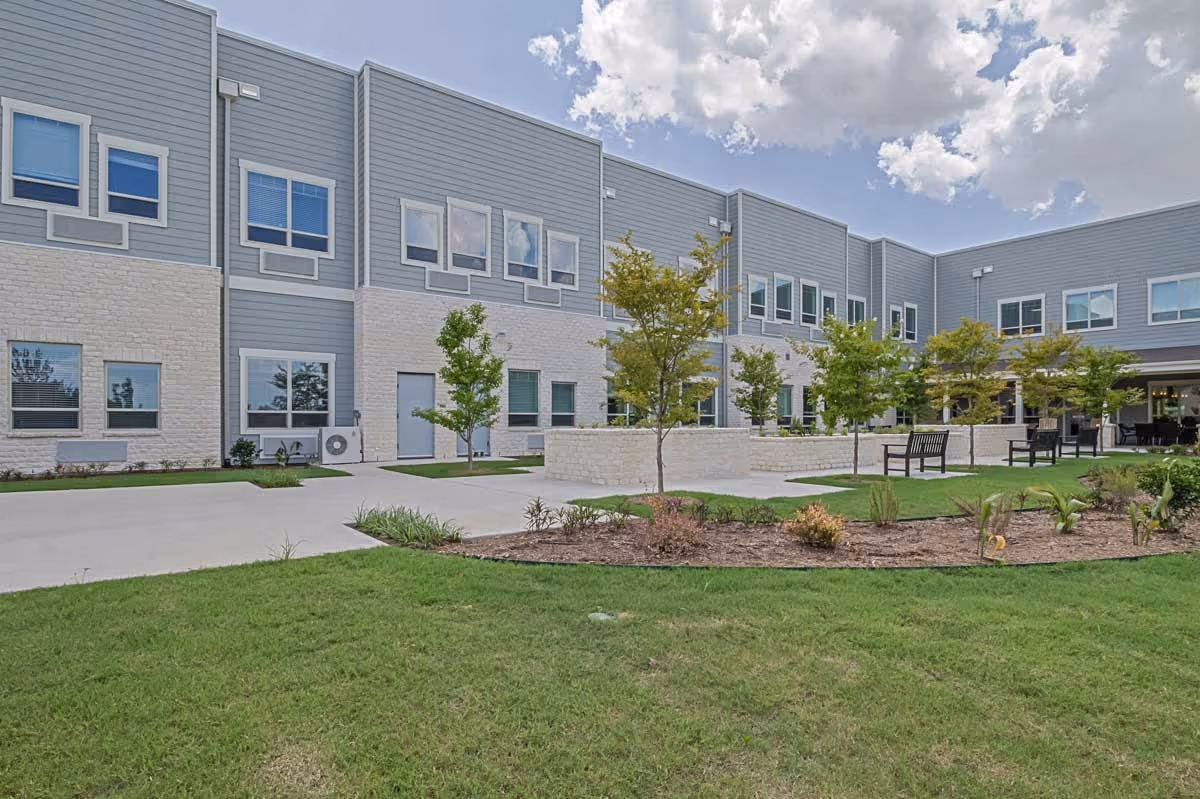 Exterior view of Midtowne Assisted Living and Memory Care building showing a two-story structure with gray siding and white stone accents. There is a landscaped garden area with small trees, shrubs, and benches on a concrete walkway under a partly cloudy sky.