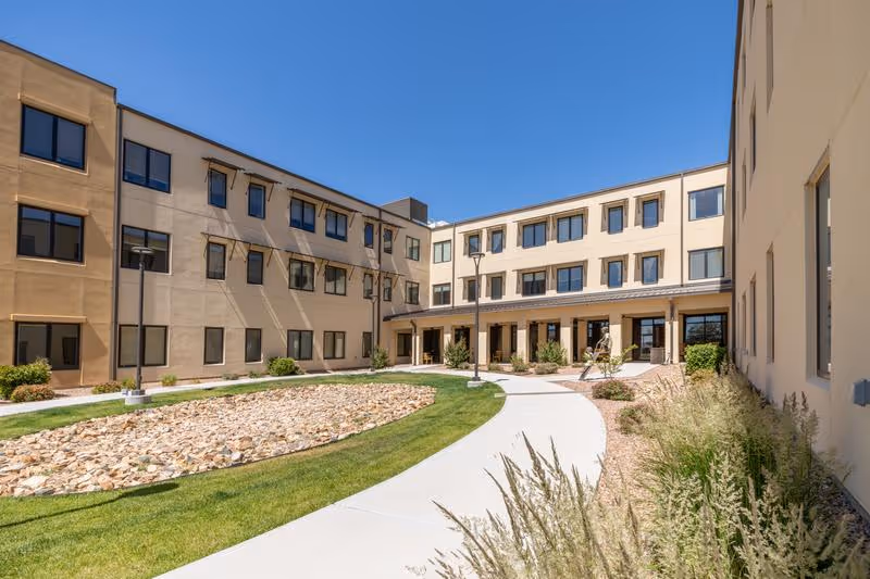 Curved walkway and landscaped courtyard surrounded by a three-story beige residential building under a clear blue sky.
