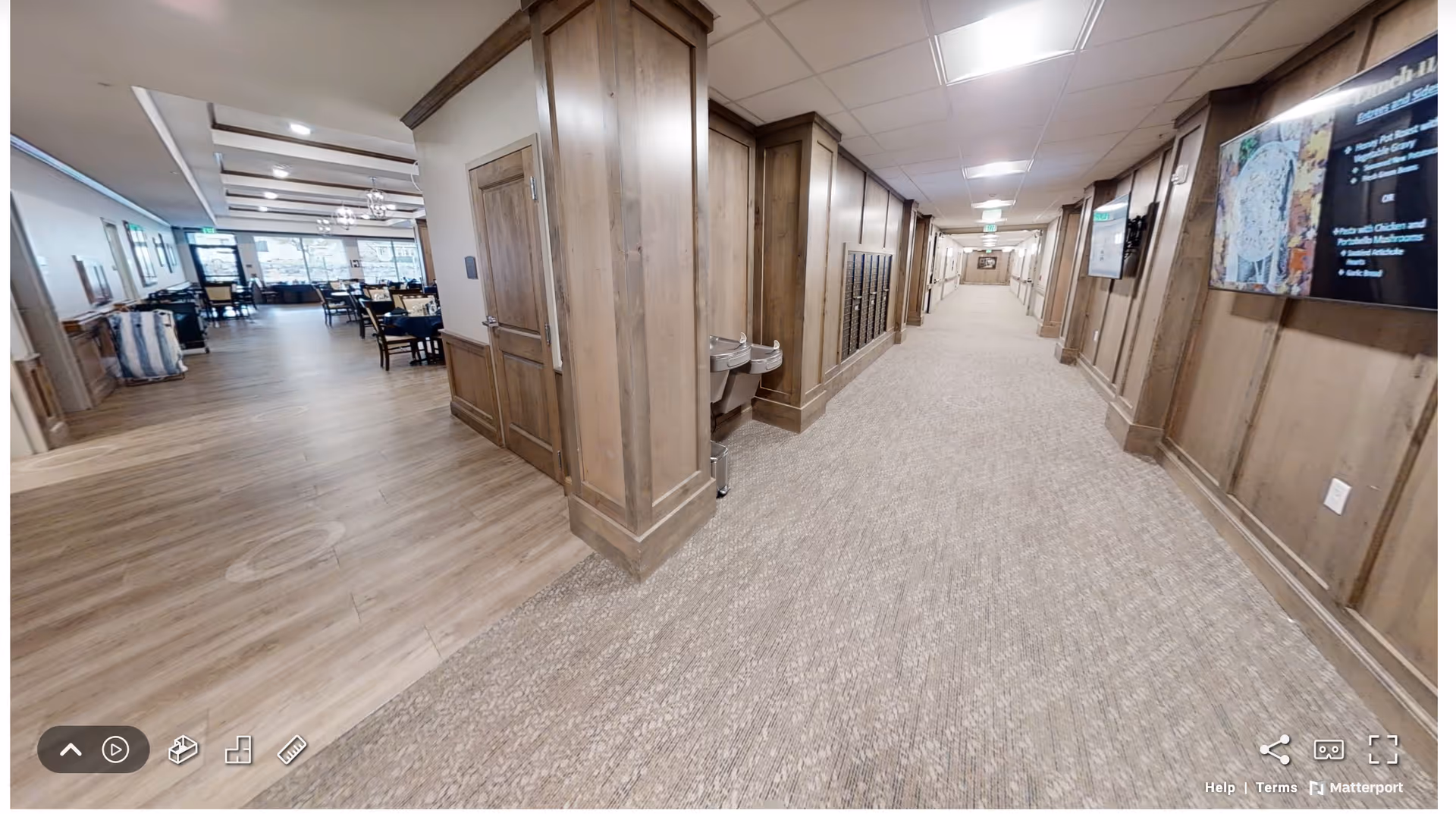 A wide hallway in an assisted living facility with carpeted flooring and wood-paneled walls. To the left, there is an open dining area with wooden floors, tables, and chairs. The hallway has a water fountain and mailboxes along the wall, with ceiling lights illuminating the space.