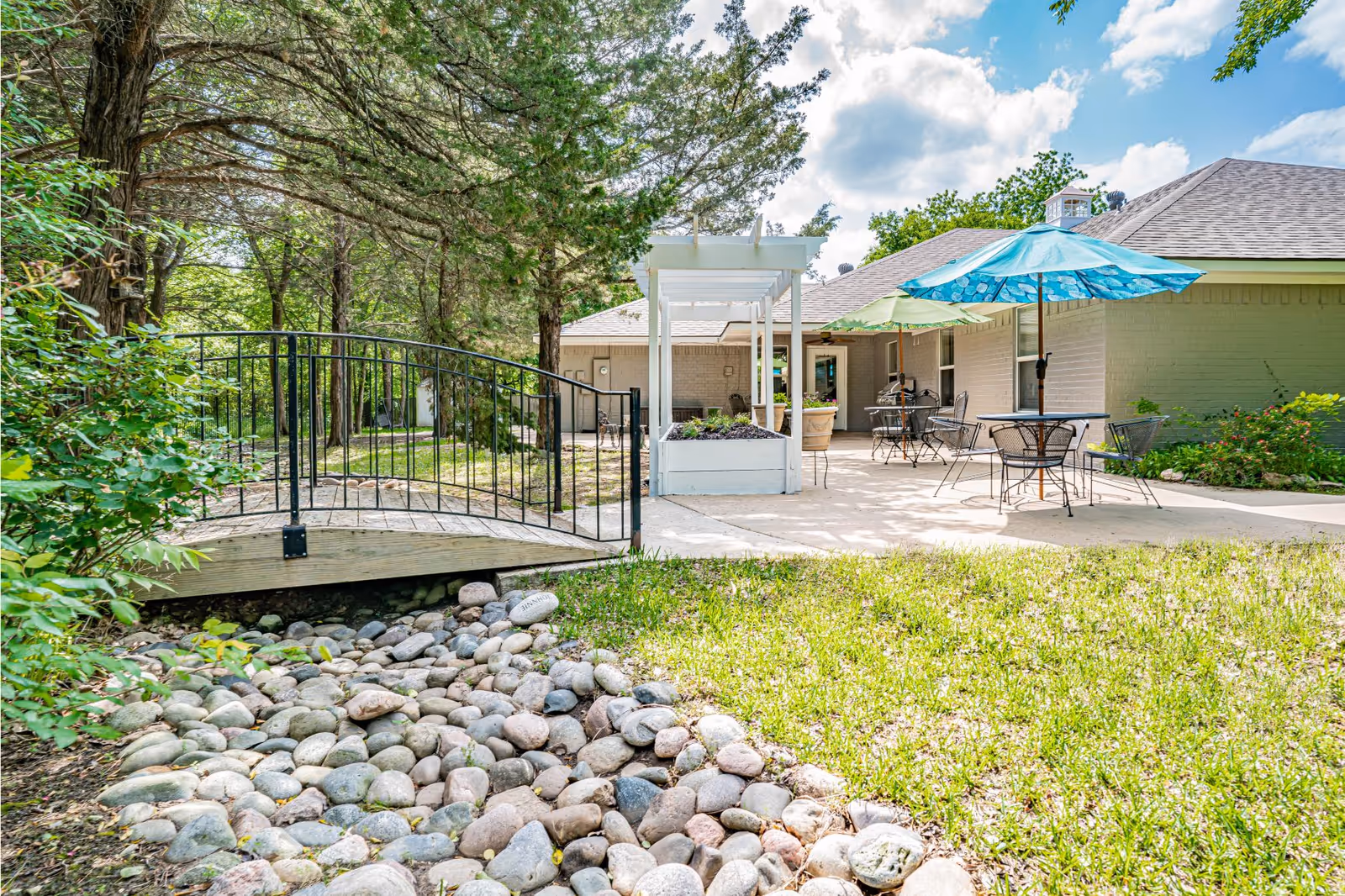 Outdoor patio area at Silver Leaf Assisted Living & Memory Care featuring a small wooden bridge over a dry rock creek, metal tables with chairs, and colorful umbrellas providing shade. The patio is adjacent to a light-colored building surrounded by trees and greenery under a partly cloudy sky.