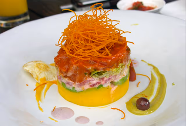 A gourmet plated dish with layered ingredients including a yellow base, green and pink middle layers, topped with a red sauce and crispy fried thin strips. The plate is garnished with a green sauce, a small dark berry, and some additional sauce drops and garnishes on a white plate.