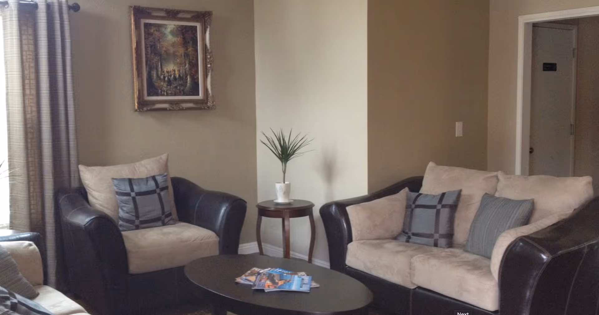 A cozy living room area with beige and dark brown cushioned armchairs and a sofa, decorated with patterned throw pillows. A small round wooden side table with a potted plant sits between the chairs. A coffee table with magazines is in the center. Beige walls, a framed painting, and a window with curtains are visible.