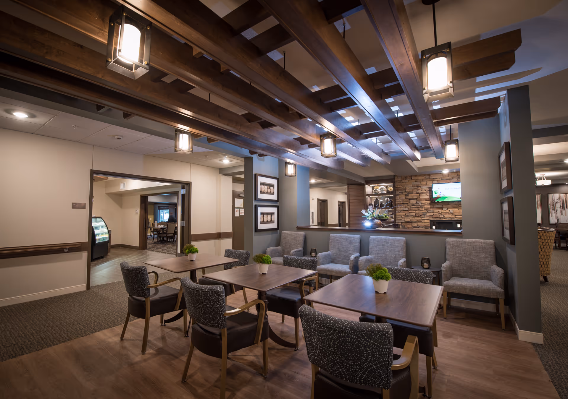 Communal dining and lounge area with tables, chairs, armchairs, and decorative wooden ceiling beams.