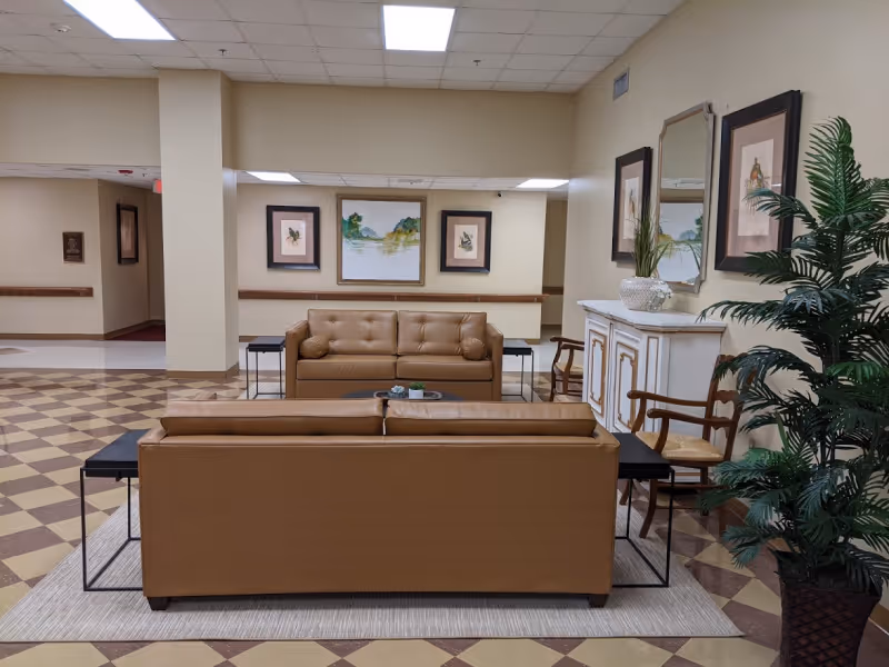 A seating area in a nursing home featuring two tan leather sofas facing each other with a small round table in between. The area is decorated with framed artwork on the wall, a white cabinet with a mirror above it, two wooden chairs, and a large potted plant. The floor has a checkered pattern and the ceiling has fluorescent lighting.
