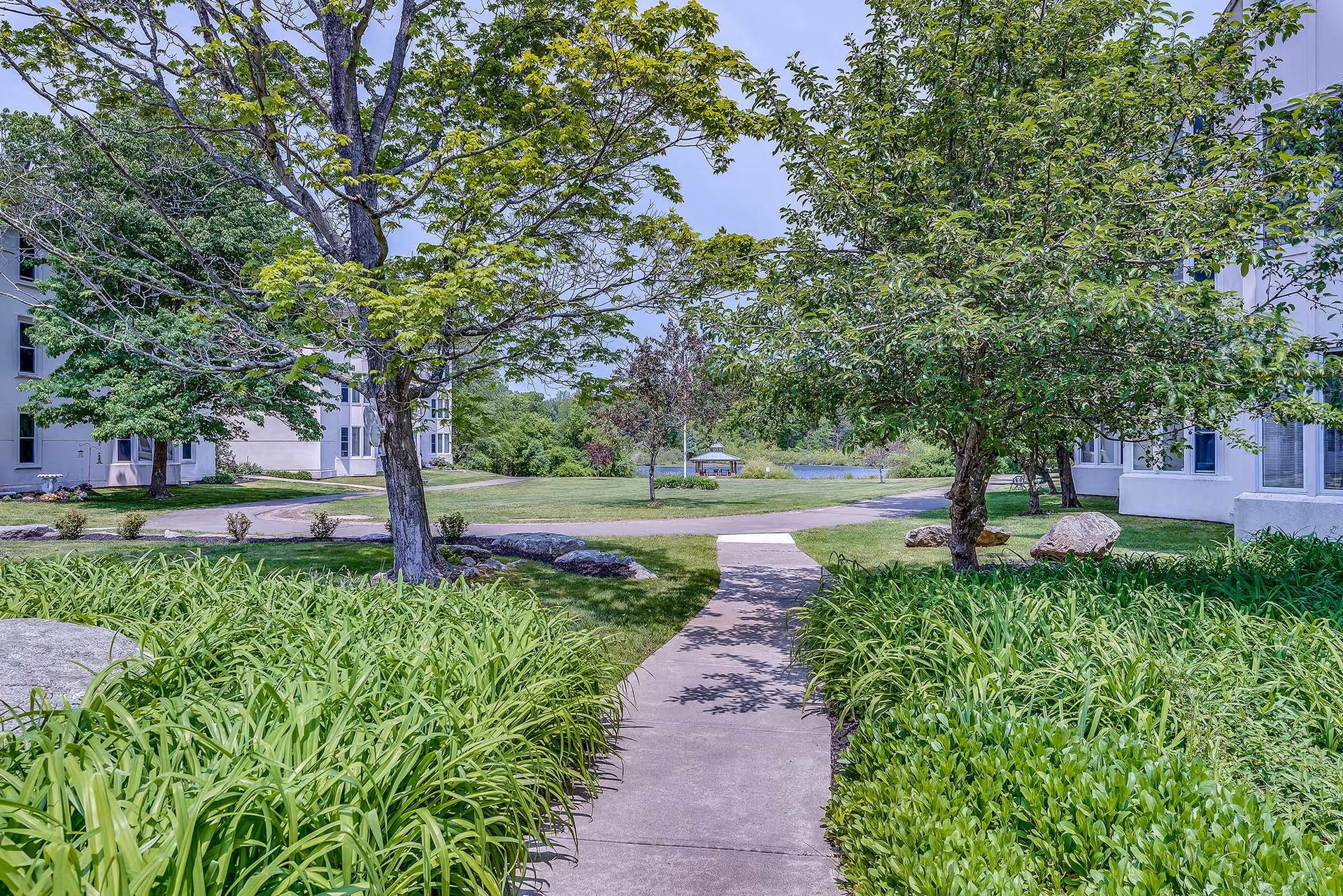Sidewalk leading through landscaped lawns and trees between retirement residence buildings toward a distant gazebo by a pond.