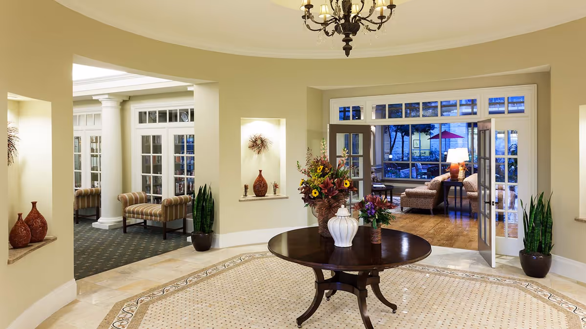 Bright elegant lobby/common area with a round table and floral centerpiece, chandelier, and seating visible through open French doors.