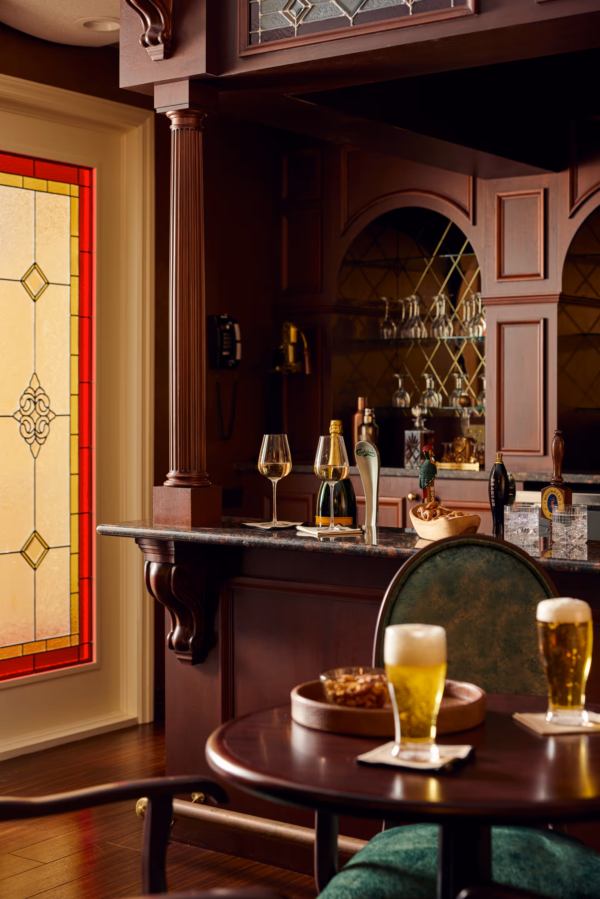 A cozy bar area with dark wood cabinetry and a stained glass window. The bar counter has two glasses of white wine, a bottle, and beer taps. In the foreground, there is a round wooden table with two glasses of beer and a bowl of snacks, accompanied by green upholstered chairs.
