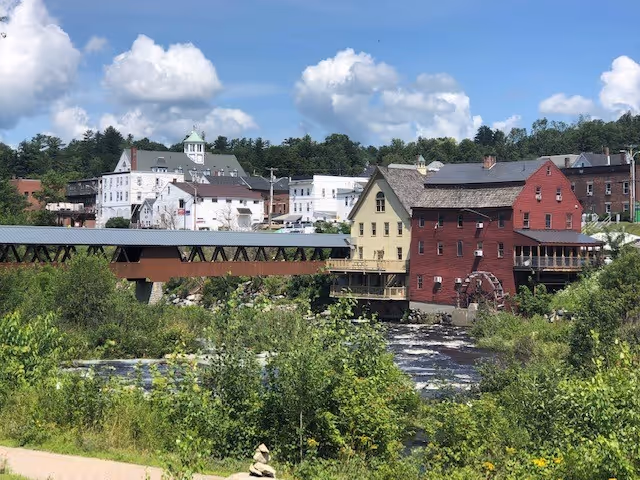 Riverfront scene showing a covered wooden bridge and red and yellow mill buildings with a waterwheel beside a flowing river and green foliage under a blue sky.