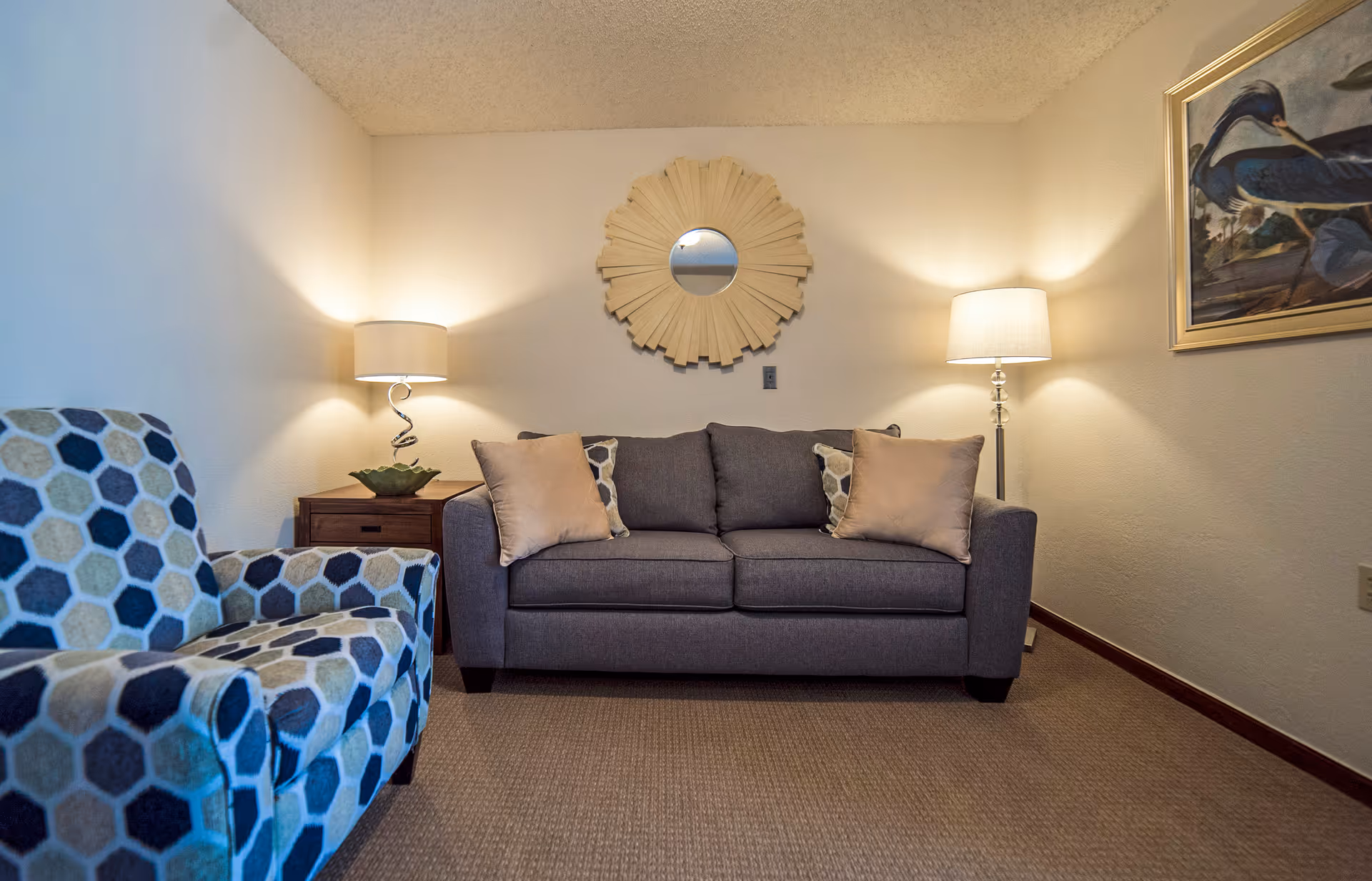 Cozy living room with a gray sofa flanked by two lamps, a patterned armchair, decorative wall mirror and artwork.