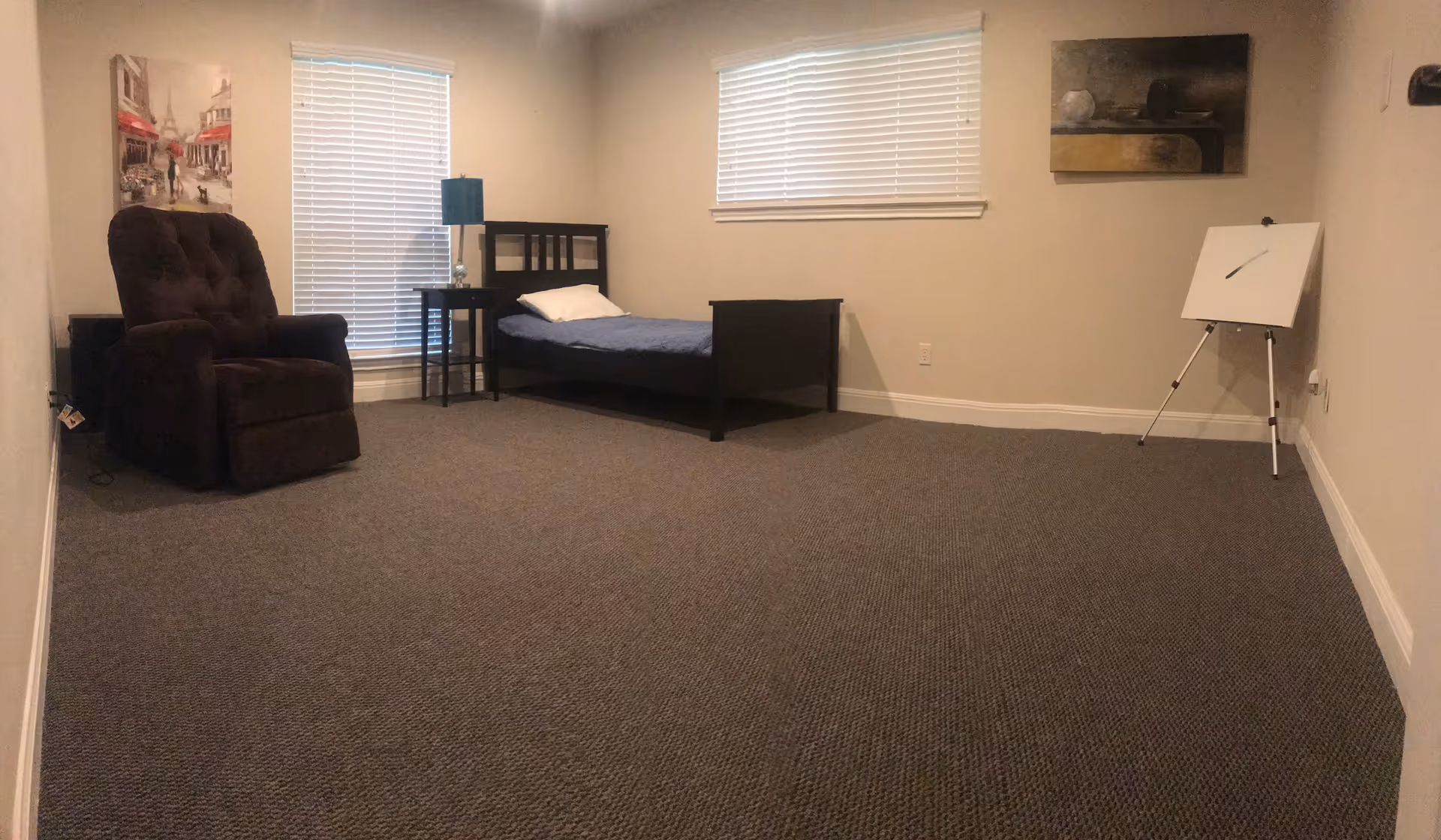 A simple bedroom with a single bed with blue bedding and a white pillow, a dark brown recliner chair, a small black side table with a blue lamp, two windows with blinds, a painting on the left wall, another painting on the right wall, and a white easel with a blank canvas in the corner.