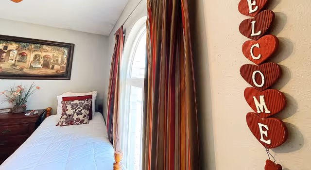 Single bed in a warmly decorated room with window, dresser, and welcome heart decor.