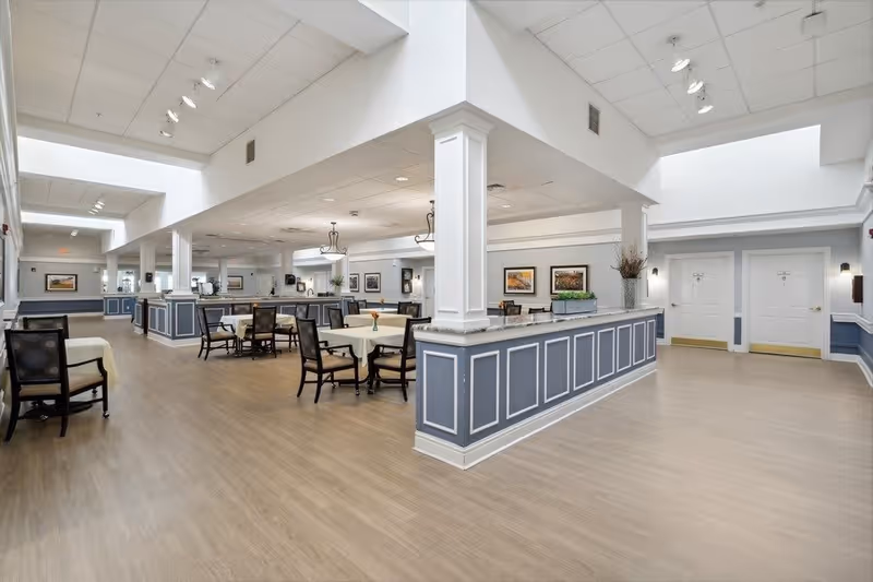 A spacious and well-lit dining area with several round tables covered with light-colored tablecloths and surrounded by dark wooden chairs. The room features light wood flooring, white walls with blue paneling, and decorative columns. There are framed pictures on the walls and ceiling lights providing illumination. Doors and additional seating areas are visible in the background.