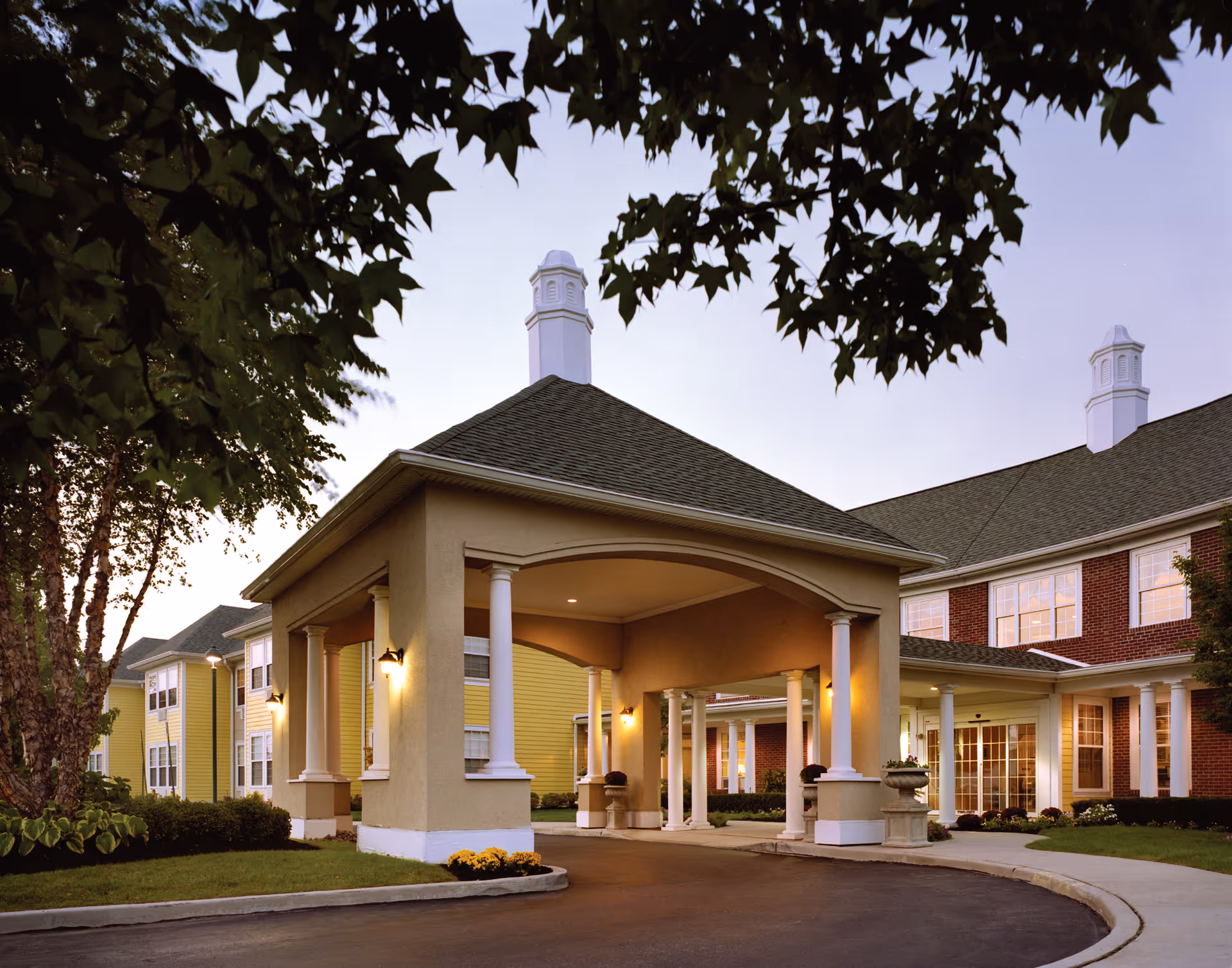 Exterior view of Brandywine Huntington Terrace by Monarch showing the entrance with a covered driveway supported by white columns, surrounded by well-maintained landscaping and trees, during dusk.