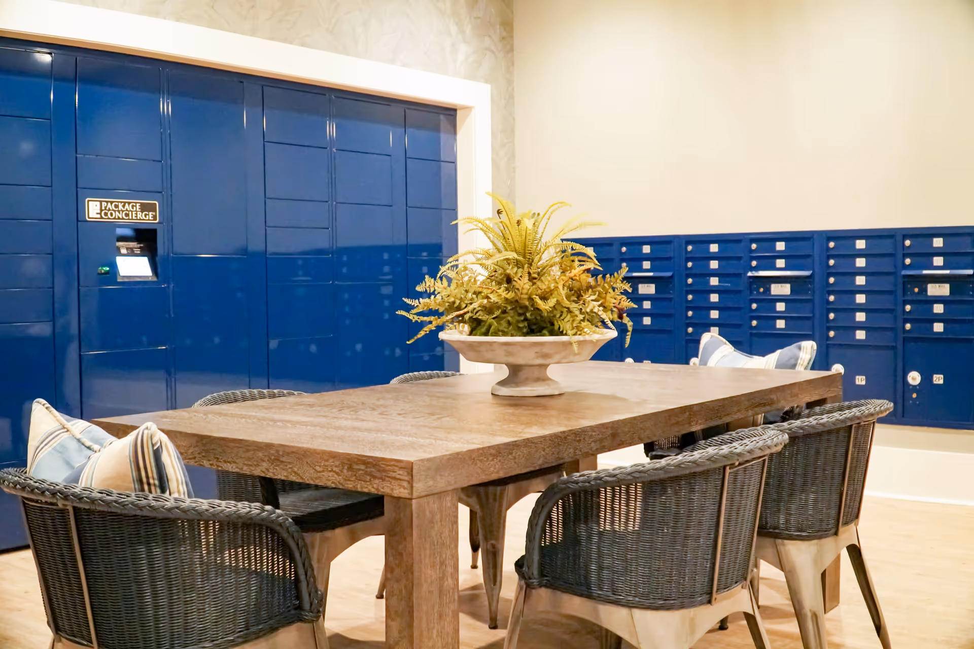 Interior room with a wooden table surrounded by wicker chairs with cushions, a decorative plant centerpiece on the table, and blue package concierge lockers and mailboxes against the walls.