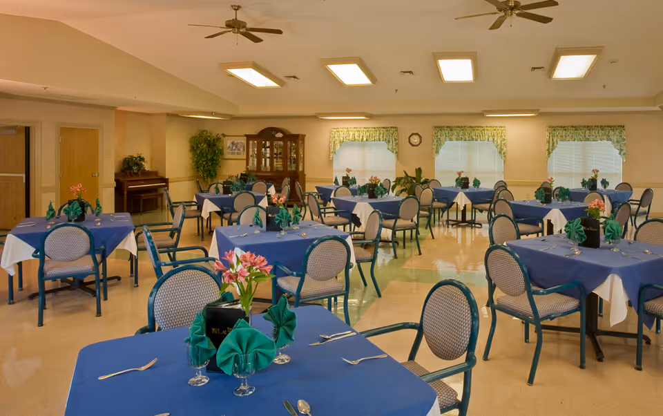 A dining room with multiple tables covered in blue tablecloths and white underlays, each set with green folded napkins, silverware, and small floral centerpieces. The room has beige walls, three windows with green floral valances, ceiling fans, and overhead fluorescent lights. There is a wooden china cabinet and a piano against the far wall.