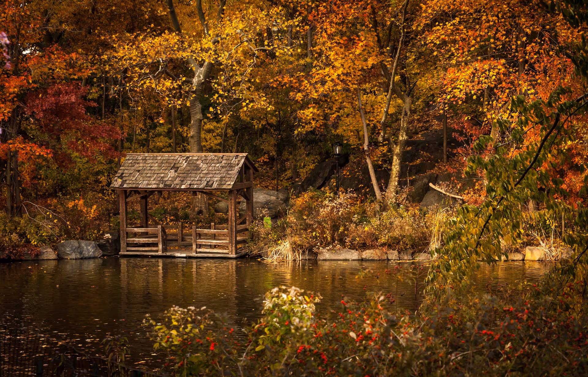 A small wooden gazebo with a shingled roof situated on the edge of a calm pond, surrounded by trees with vibrant autumn foliage in shades of yellow, orange, and red.