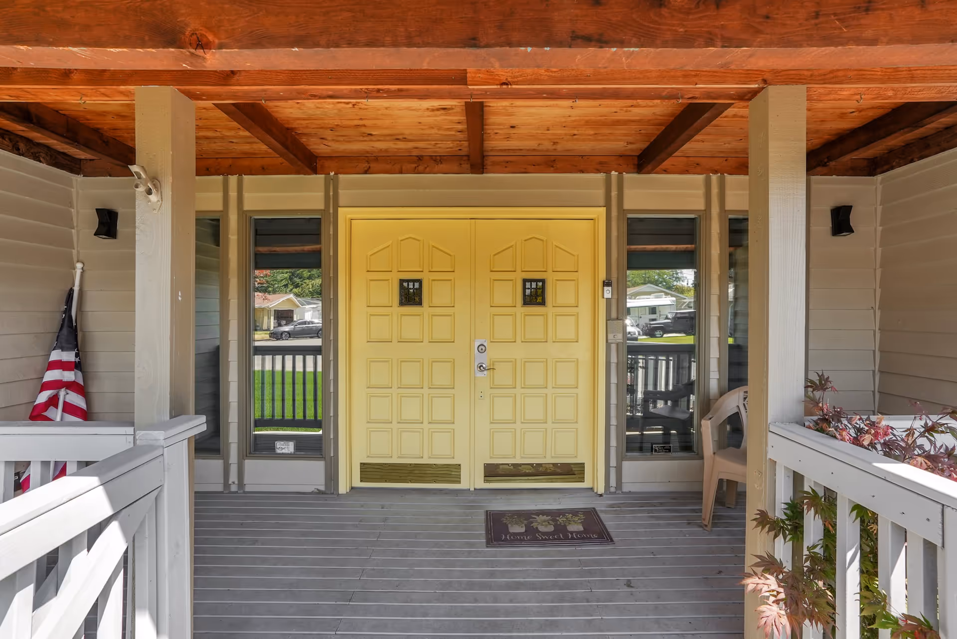 Front porch of a building with a wooden ceiling and gray floorboards. There is a double yellow door in the center with small square windows near the top. On the floor in front of the door is a mat that reads 'Home Sweet Home'. To the left of the door is an American flag, and to the right is a beige plastic chair and some plants. Two narrow windows flank the door, showing a view of the street and houses outside.