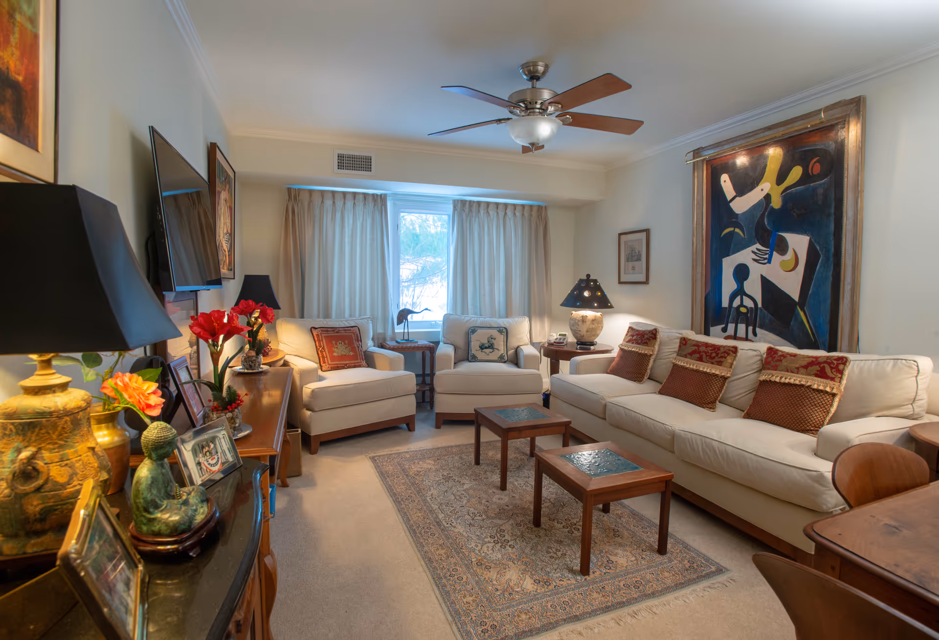 A cozy living room with a beige sofa adorned with red and brown decorative pillows, two matching armchairs, a patterned area rug, two small wooden coffee tables with glass tops, and a ceiling fan with wooden blades. The room features a large abstract painting on the wall, a flat-screen TV mounted on the opposite wall, and various decorative items including lamps, flowers, and framed photos on a wooden sideboard. A window with beige curtains lets in natural light.