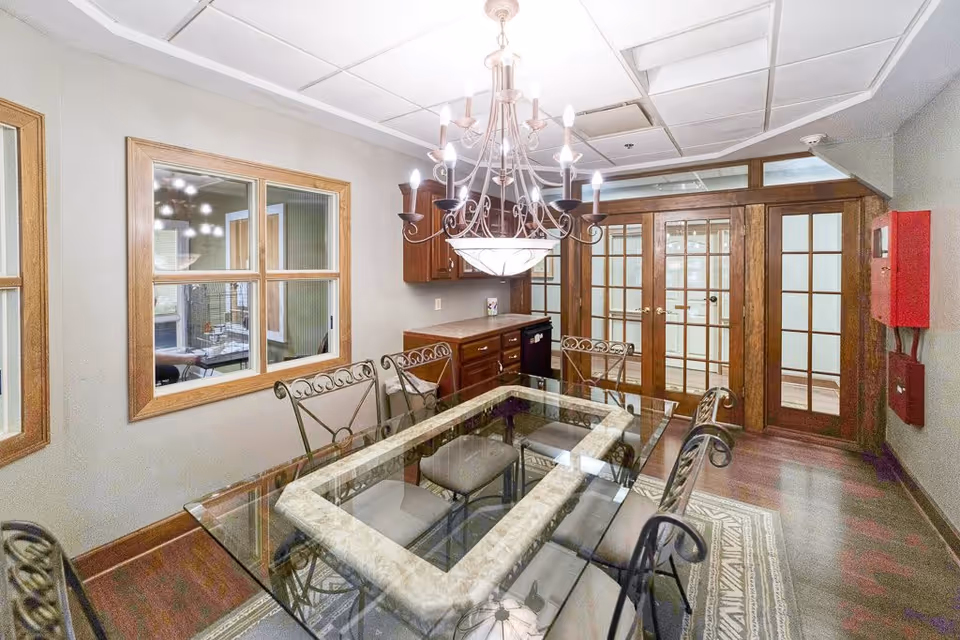 A dining room with a glass-top table surrounded by six metal chairs with cushions. Above the table hangs an ornate chandelier with multiple lights. The room features wooden framed windows and French doors with glass panels. There is a small cabinet with a countertop and a dishwasher underneath on one side of the room. The floor has a patterned rug and wooden flooring.