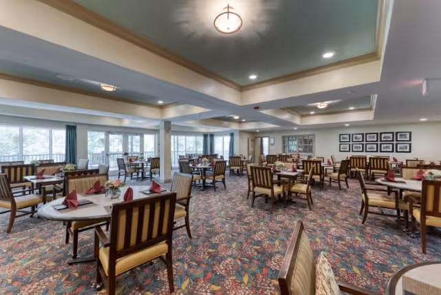 Spacious dining room with multiple round tables and wooden chairs with cushioned seats and backs. Each table is set with folded red napkins and small floral centerpieces. Large windows along one wall allow natural light to fill the room, and the ceiling features recessed lighting and a decorative central light fixture. The carpet has a colorful floral pattern, and framed pictures hang on one wall.