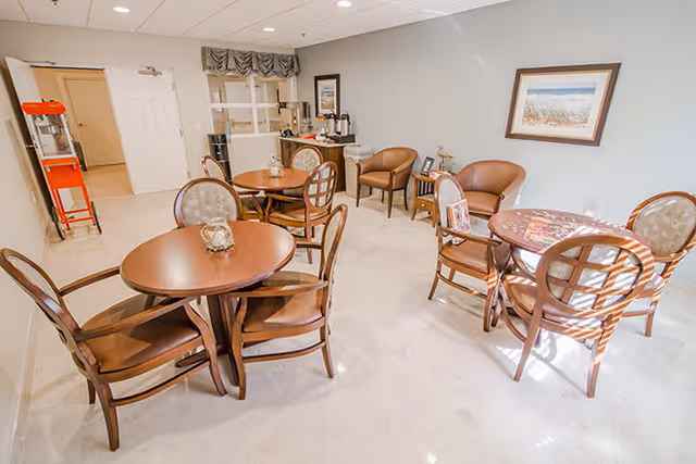A bright and clean common area with multiple round wooden tables and chairs arranged for seating. There are two brown armchairs against the wall, a small side table with a lamp, and a framed picture hanging on the wall. A coffee station with supplies is visible in the corner near a door. The floor is light-colored tile, and the walls are painted a soft gray.
