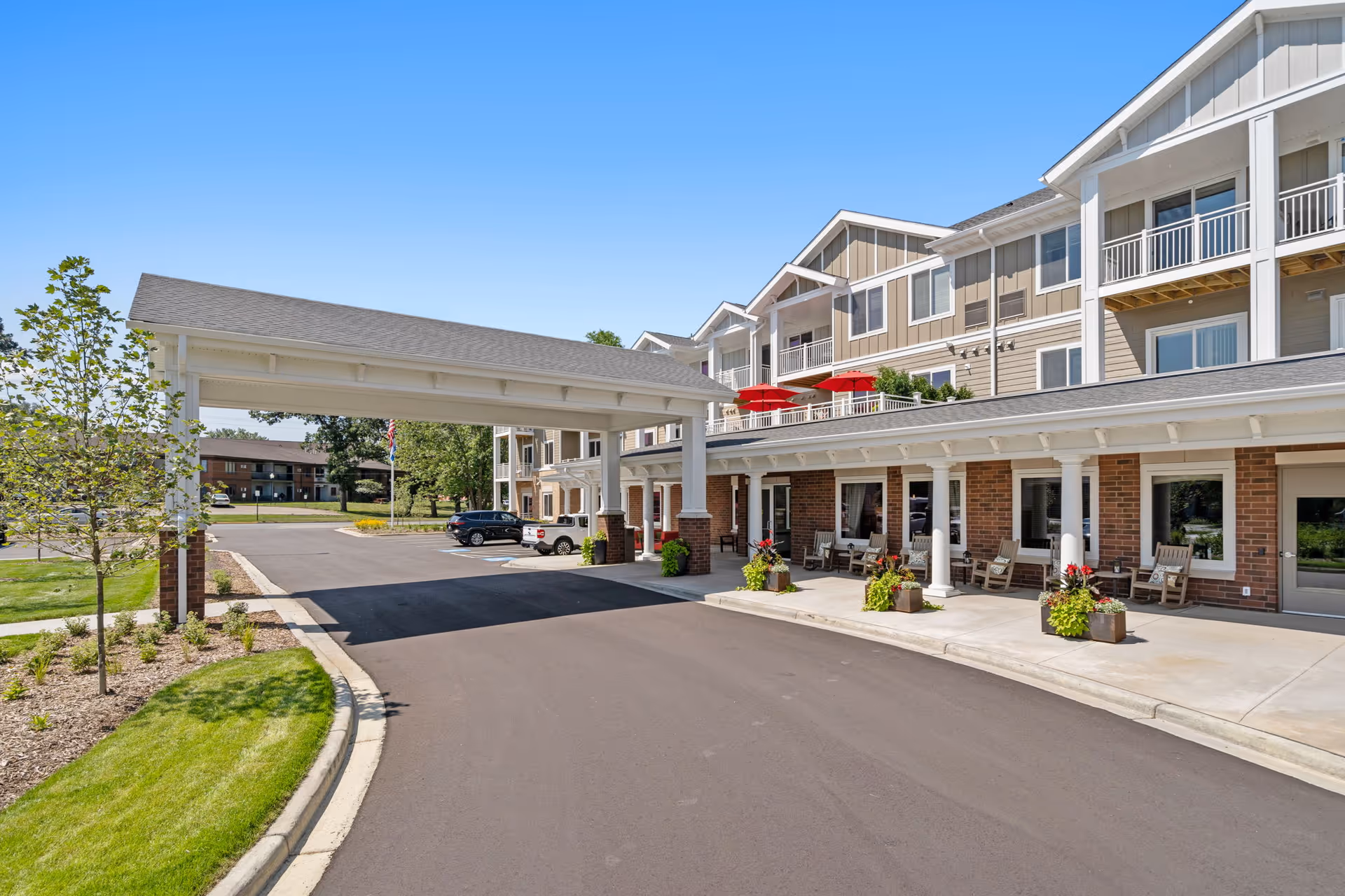 Exterior view of The Sycamore of River Falls senior living facility showing a covered entrance driveway, landscaped greenery, parked cars, and a multi-story building with balconies and outdoor seating areas under a clear blue sky.