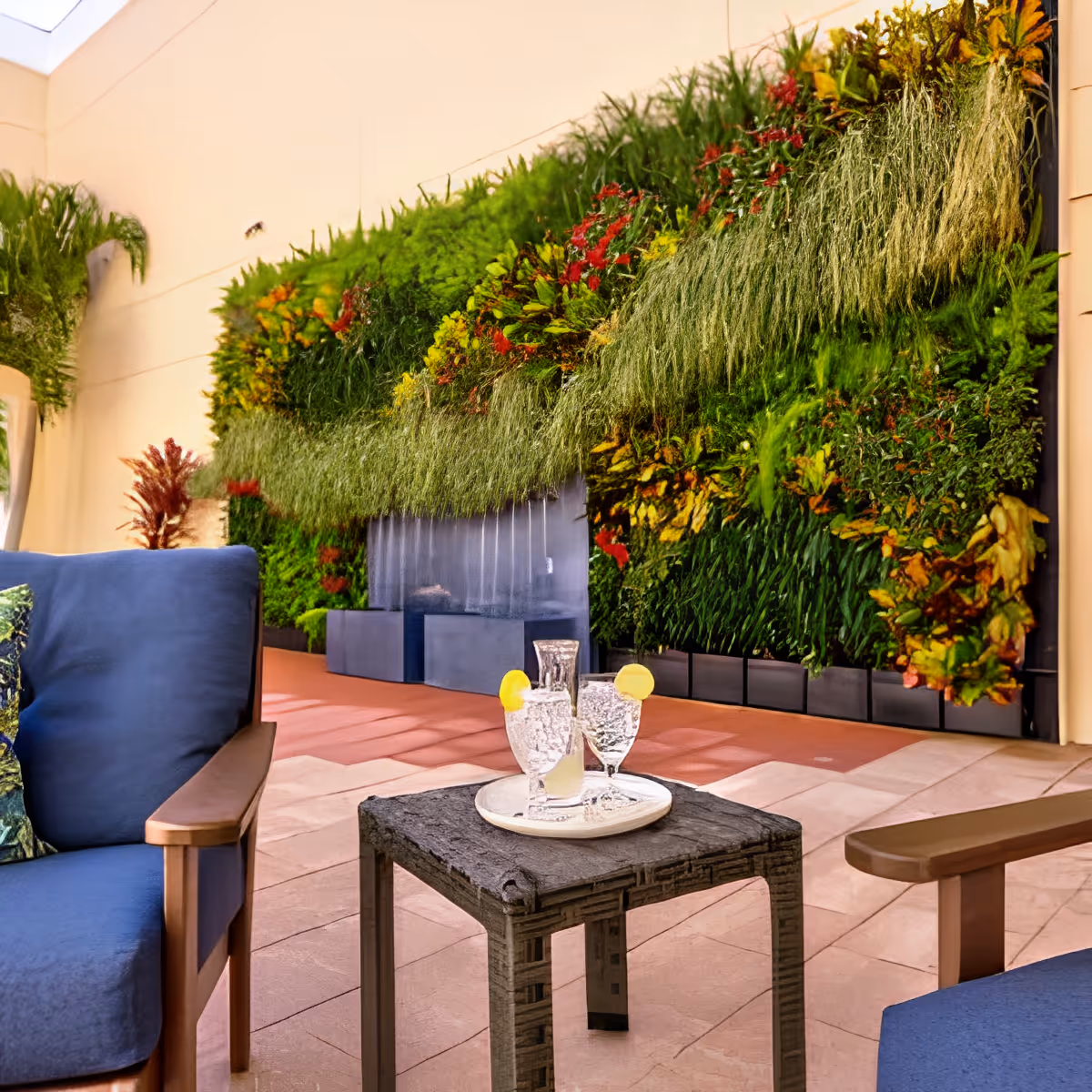 Outdoor seating area with two blue cushioned chairs and a small table holding a tray with a pitcher and two glasses of water garnished with lemon slices. Behind the seating area is a vertical garden wall with various green and colorful plants.