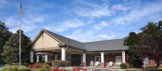 Exterior view of a single-story brick building with a covered entrance, white columns, and a well-maintained garden with red and white flowers in front. An American flag is flying on a flagpole to the left of the building under a partly cloudy sky.