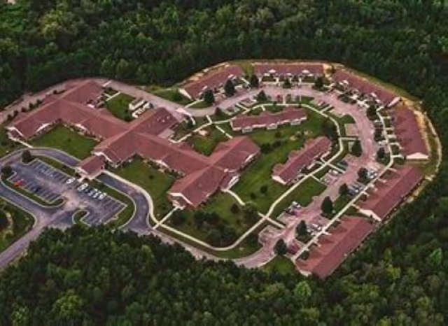 Aerial view of a senior living campus with multiple connected red-roof buildings, driveways, parking areas, and surrounding trees.