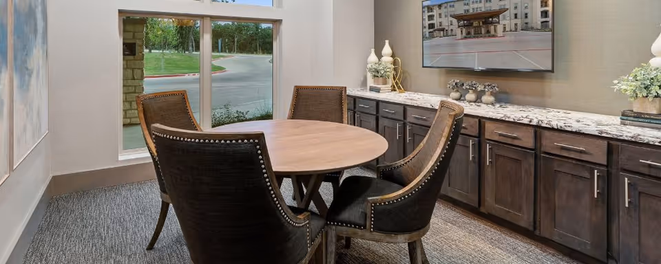 Round wooden table with four upholstered chairs in a small dining area featuring dark wood cabinetry, a marble countertop, wall-mounted TV, and a window overlooking landscaping.