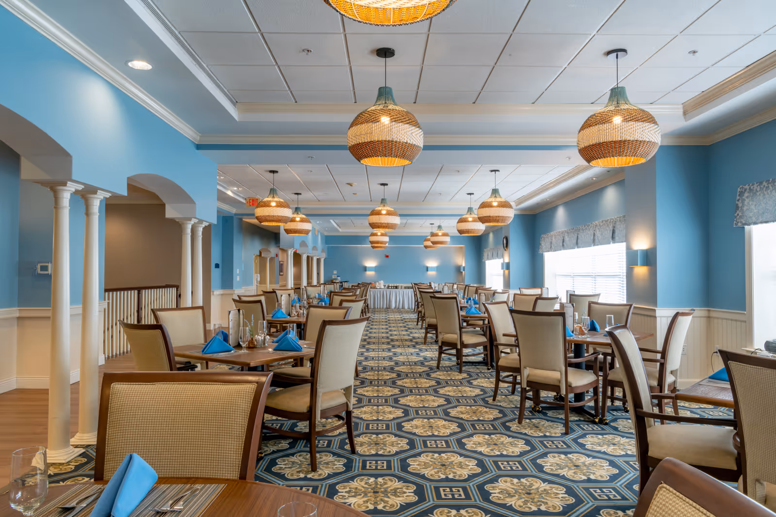 A spacious dining room with blue walls and patterned carpet featuring multiple tables set with blue napkins, glasses, and silverware. The room is illuminated by several hanging woven pendant lights and has large windows with valances letting in natural light. White columns and arches line one side of the room.