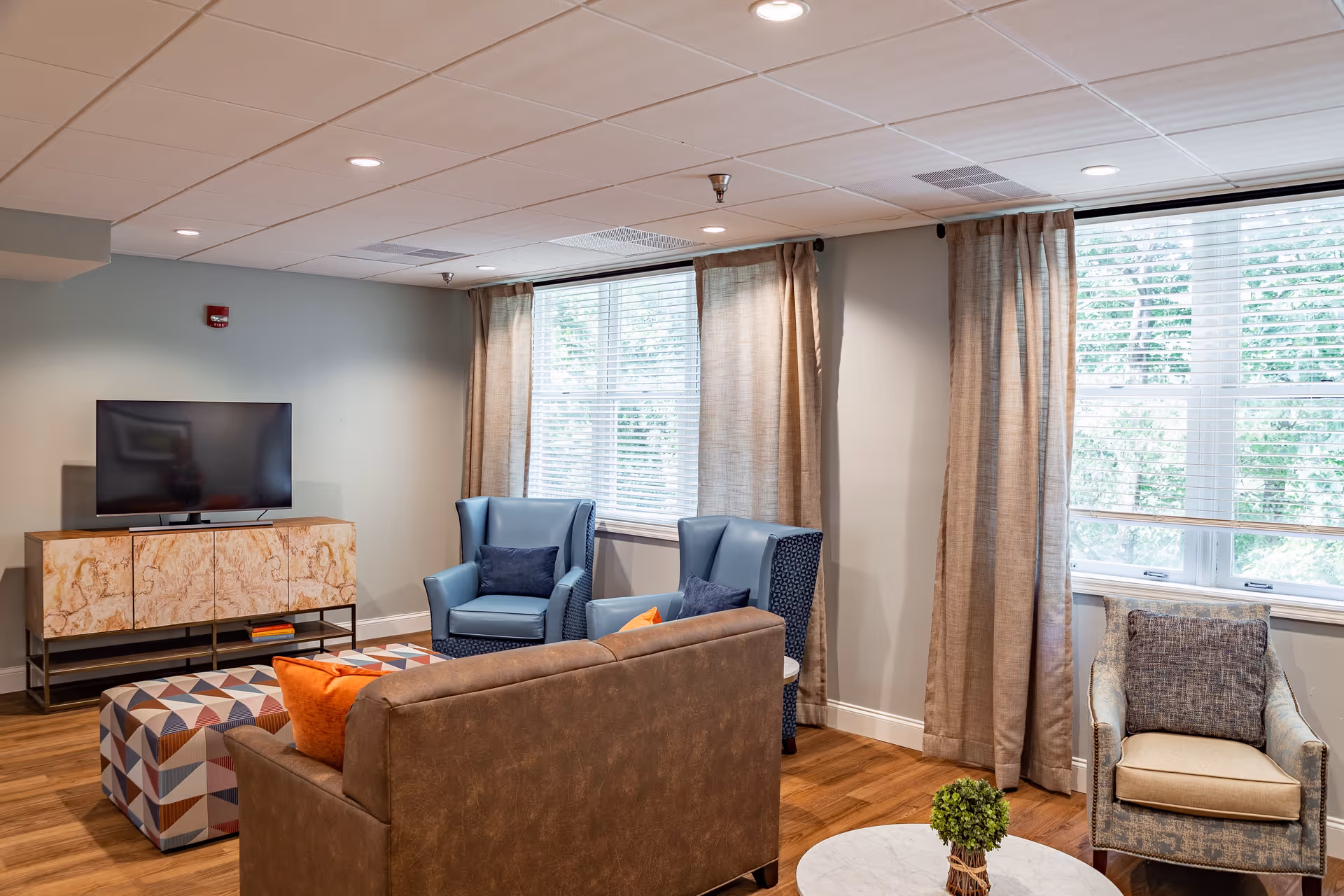 A cozy living room with a brown sofa, two blue armchairs, a patterned ottoman, and a single beige armchair. The room has large windows with beige curtains, a flat-screen TV on a wooden stand, and a small round table with a green plant. The floor is wooden and the walls are painted light gray.