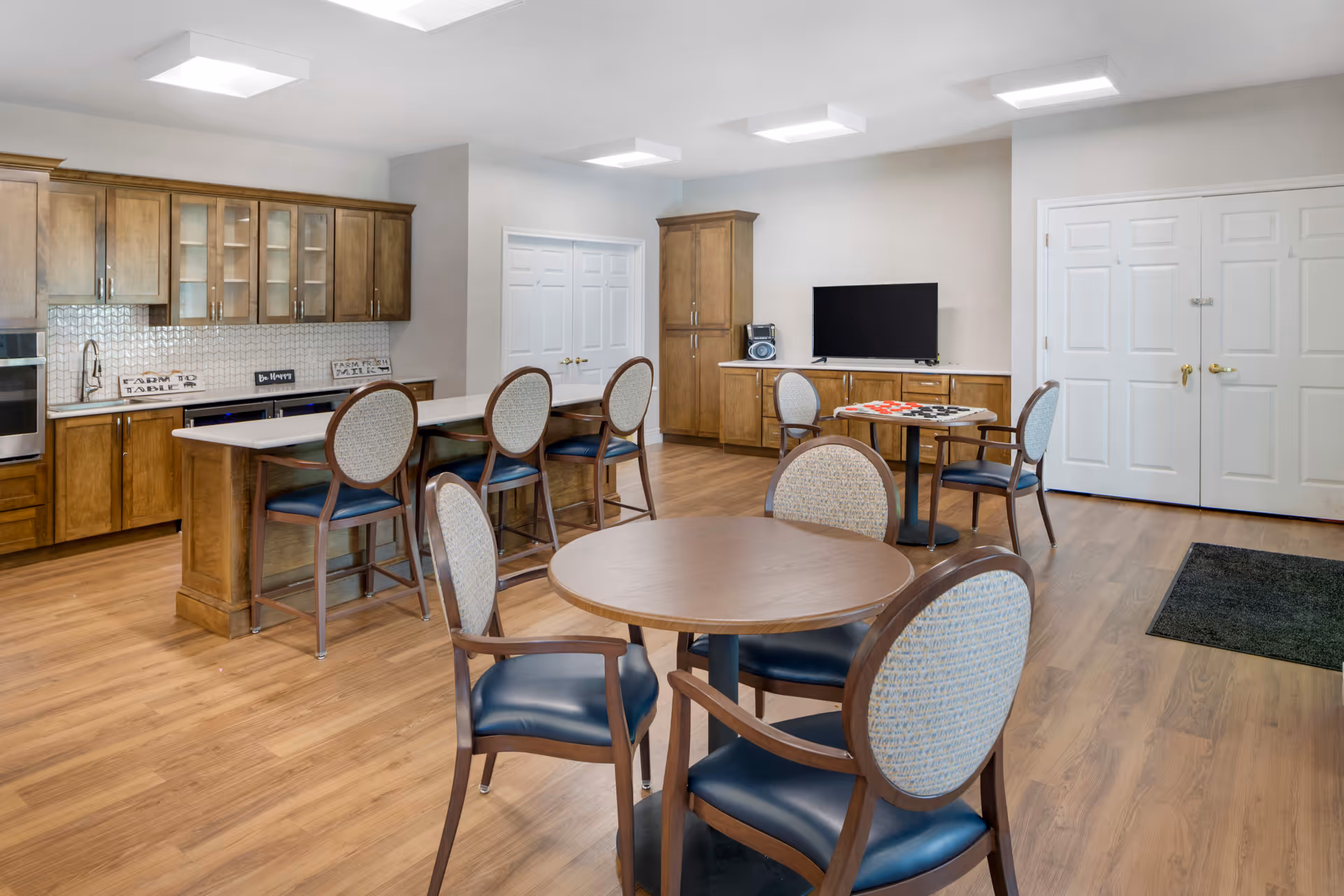 A spacious senior living common area featuring a kitchen with wooden cabinets, a long island with four chairs, and a dining area with round tables and cushioned chairs. The room has wood flooring, white walls, and ceiling lights. A flat-screen TV and a radio are placed on a cabinet against the far wall.