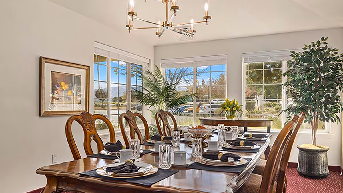Sunlit dining room with a long wooden table set for a meal and chairs in front of large windows.