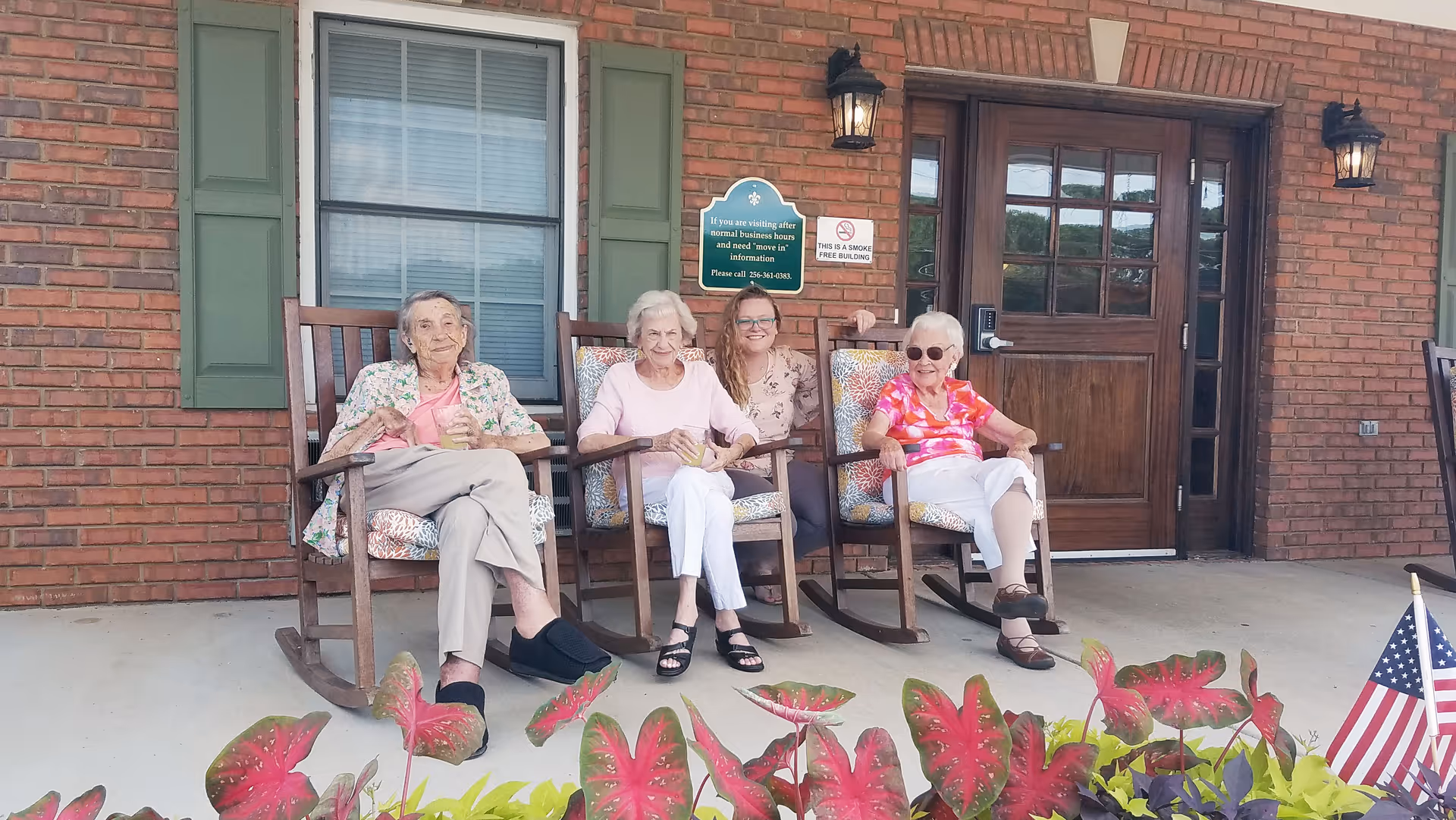 Three elderly women and a younger woman sitting on rocking chairs on a porch in front of a brick building with green shutters and a wooden door. There are colorful plants in the foreground and an American flag on the right side.