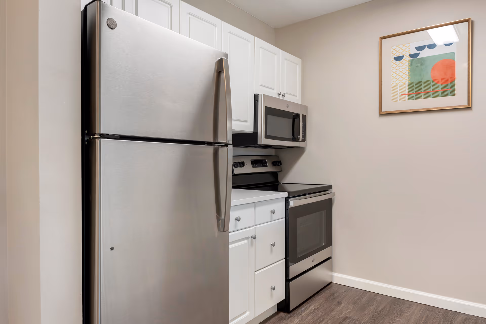 A modern kitchen area featuring a stainless steel refrigerator, a stainless steel microwave mounted above a stainless steel electric stove, white cabinetry with silver knobs, and a framed abstract artwork on a beige wall. The floor is a dark wood laminate.