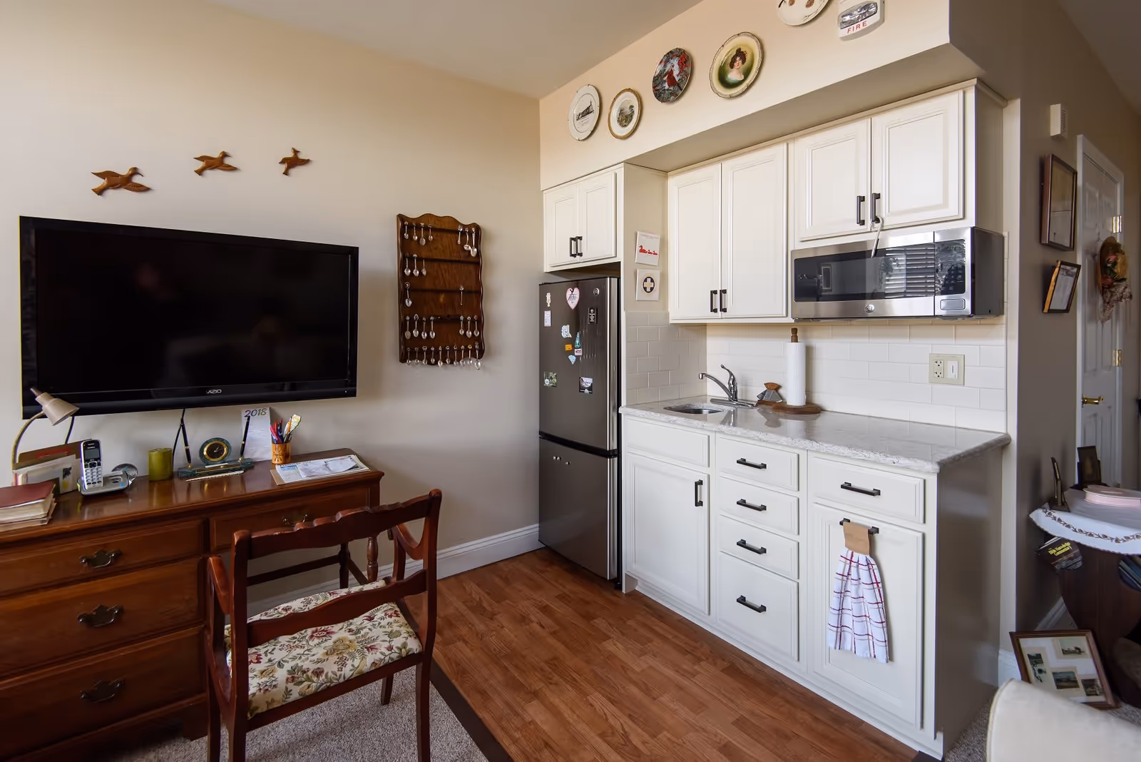 A small living space featuring a kitchenette with white cabinets, a stainless steel microwave, and a small sink. Next to the kitchenette is a stainless steel refrigerator adorned with magnets. On the left side, there is a wooden desk with a floral cushioned chair, a desk lamp, a phone, and various office supplies. Above the desk is a mounted flat-screen TV and decorative wooden birds on the wall. The floor is a combination of wood and carpet.