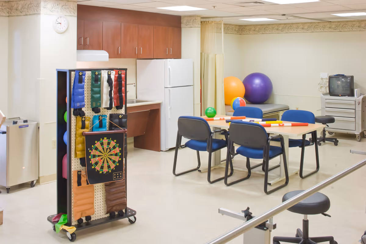 A therapy or activity room with exercise and rehabilitation equipment including colorful therapy bands, a dartboard, large exercise balls, a table with chairs, and a small TV on a cart. The room has a kitchenette area with a refrigerator and sink.