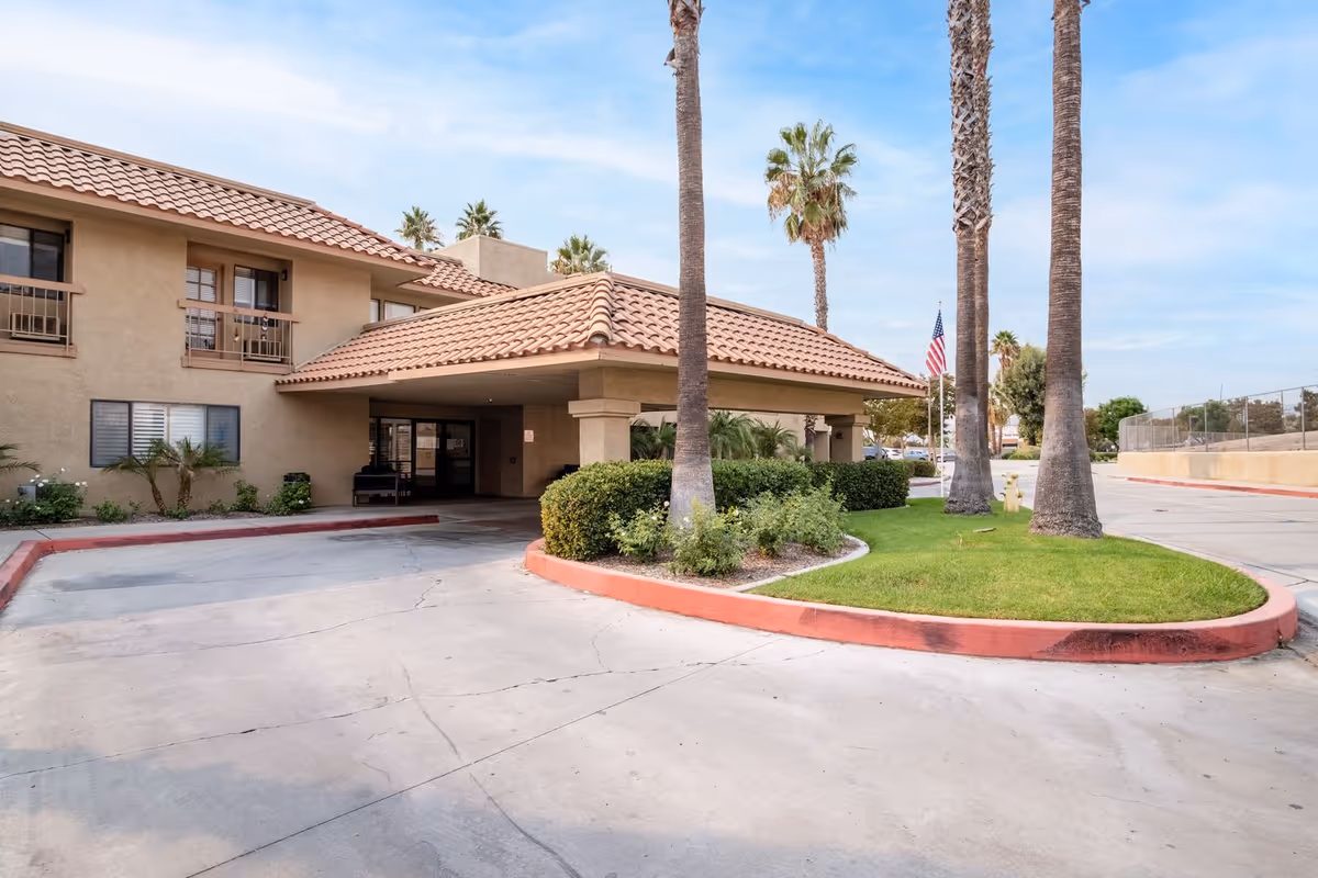 Exterior view of Vista Blue Mountain Assisted Living & Memory Care building with a covered entrance, palm trees, green lawn, and an American flag in the background under a partly cloudy sky.