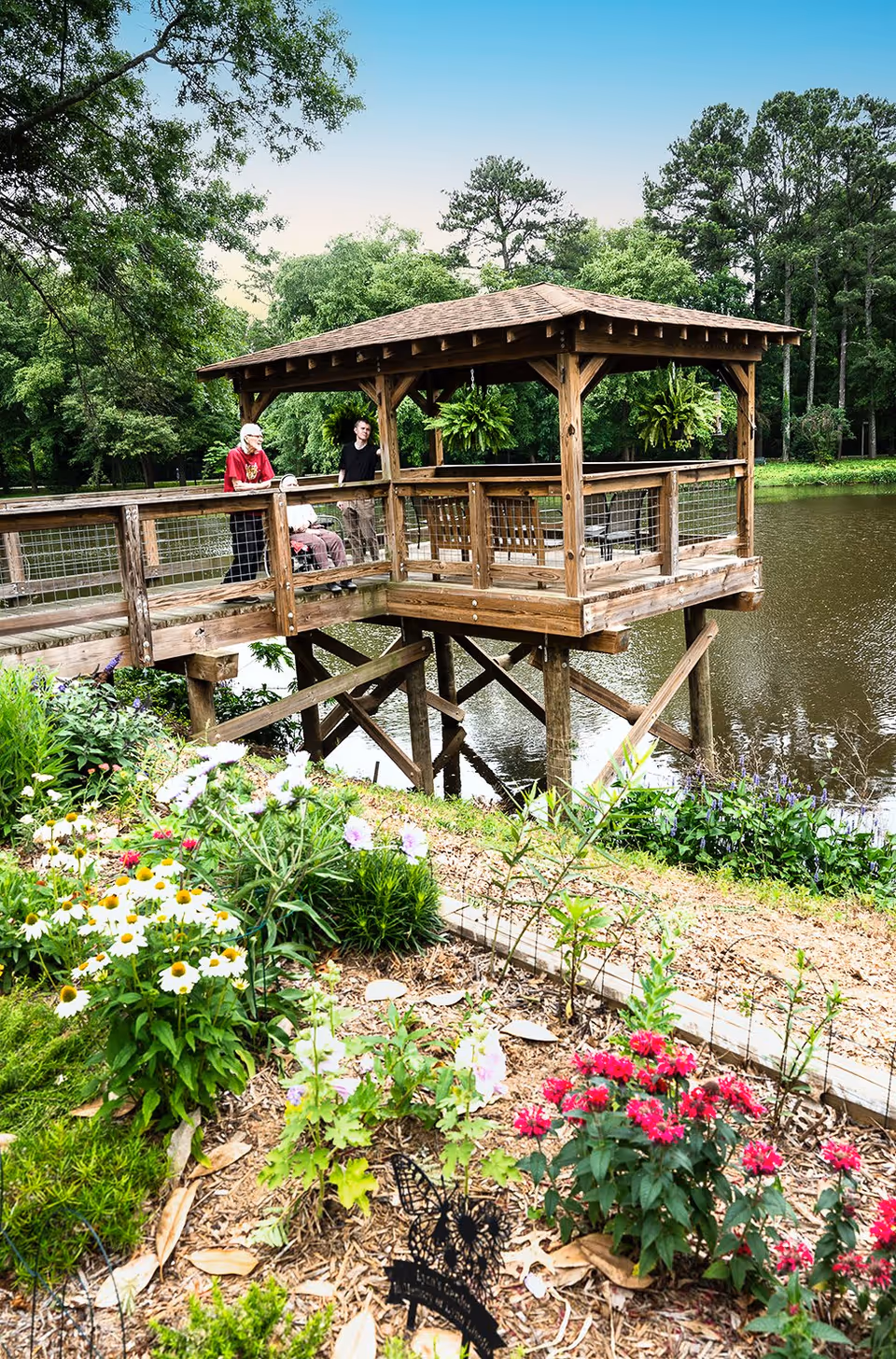 A wooden gazebo on stilts extends over a pond with a railing and wire mesh. Three people, including one in a wheelchair, are on the gazebo enjoying the view. The foreground features a garden with colorful flowers and greenery, and tall trees surround the pond under a clear blue sky.
