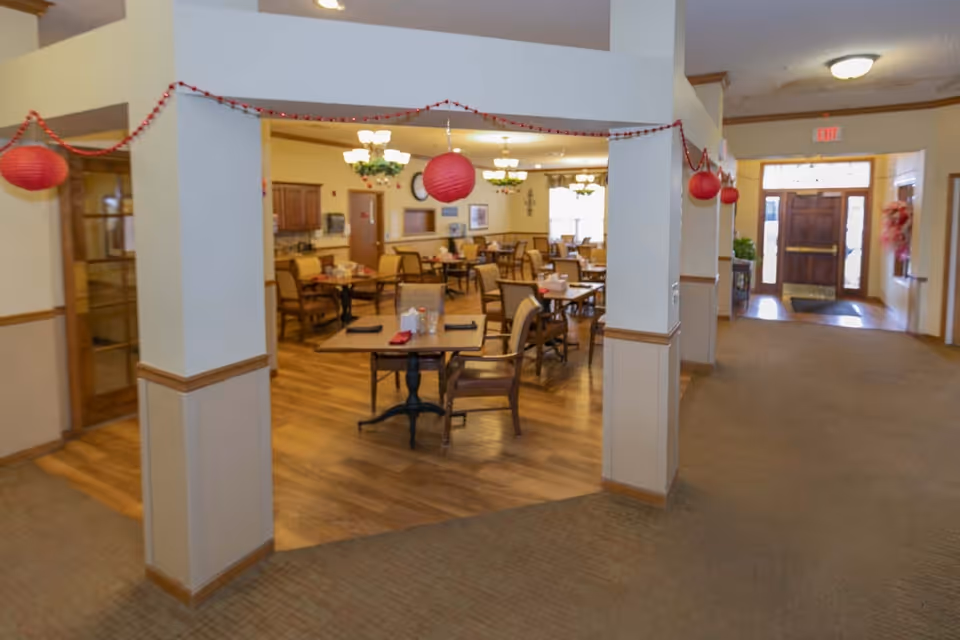 Interior view of a senior living facility dining room with multiple tables and chairs arranged neatly. The room features wooden flooring, beige walls with wooden trim, and is decorated with red hanging lanterns and garlands. There are chandeliers hanging from the ceiling and a clock on the far wall. To the right, there is a carpeted hallway leading to a wooden entrance door with glass panels.