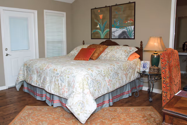 A bedroom with a large bed covered in a floral patterned bedspread and several pillows. There is a nightstand with a lamp, a framed photo, and a decorative box next to the bed. Two framed floral paintings hang on the wall above the bed. A door and window with blinds are visible on the left side, and a patterned rug covers part of the wooden floor.