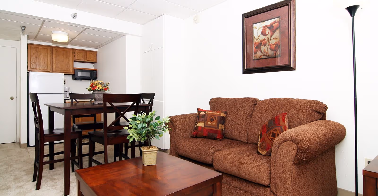 Cozy living area with a brown sofa, wooden coffee table and dining set adjacent to a small kitchen with refrigerator and cabinets.