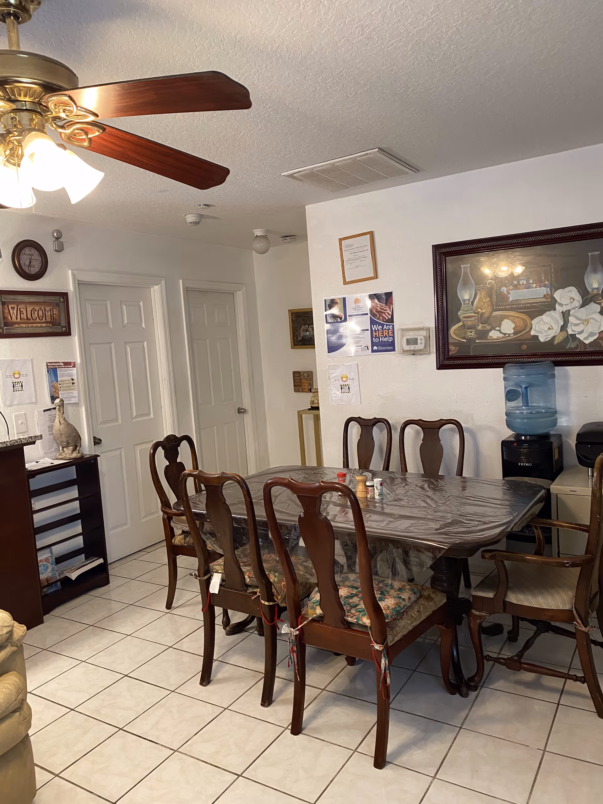Indoor dining area with a wooden table covered in plastic, six chairs, a ceiling fan, wall art, and a water cooler.