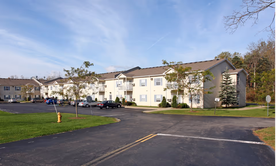 Exterior view of a two-story senior apartment building with beige siding and multiple windows. There are several small trees and a parking lot with cars in front of the building under a partly cloudy blue sky.