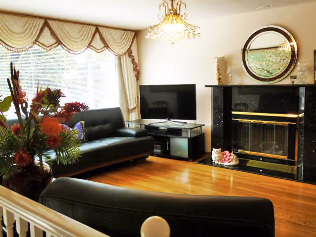 A cozy living room with two black leather sofas, a flat-screen TV on a stand, a black marble fireplace with a round mirror above it, and a large window with elegant drapes. There is a floral arrangement on the left side and hardwood flooring throughout the room.