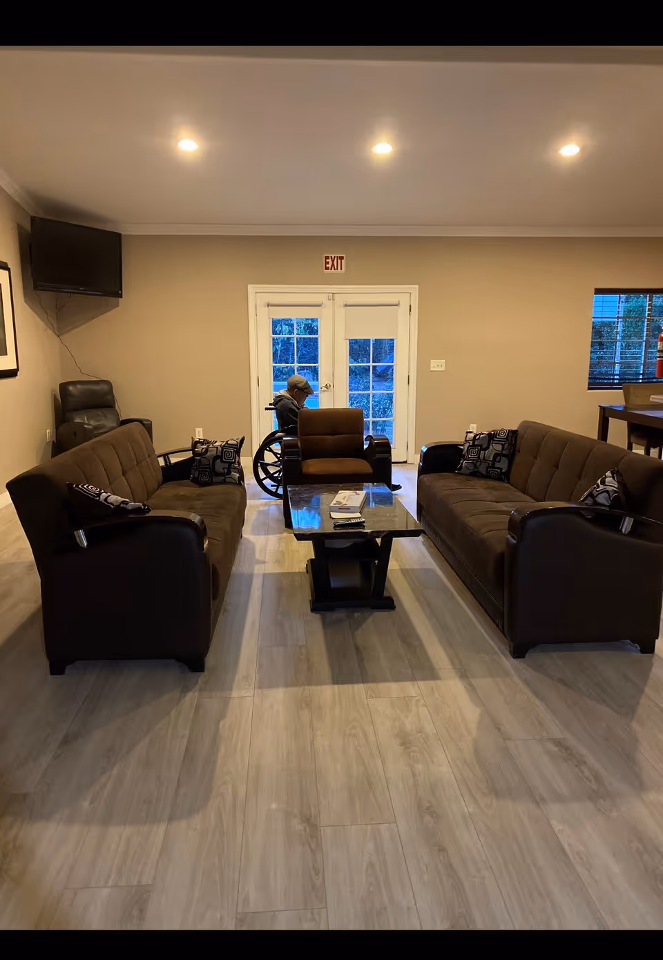 A cozy living room with two brown sofas facing each other and a matching armchair in the center. A glass-top coffee table is placed between the sofas. A person in a wheelchair is positioned near the glass double doors at the back of the room. The room has light wood flooring, beige walls, recessed ceiling lights, a wall-mounted TV in the corner, and a window with blinds on the right side.