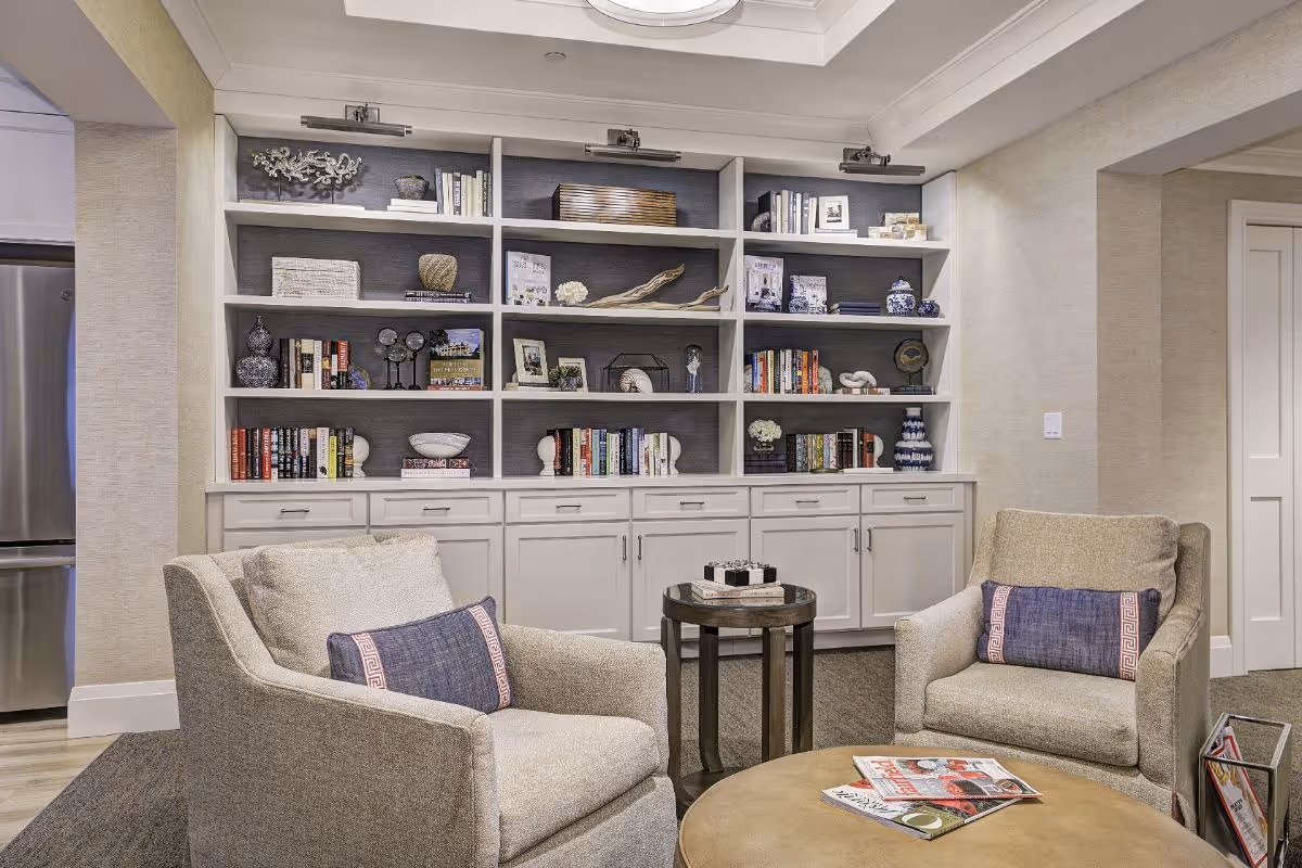 A cozy living room area with two beige armchairs, each with a blue decorative pillow, positioned around a round coffee table with magazines on it. Behind the chairs is a large built-in white bookshelf filled with books, decorative items, and artwork. The room has neutral-colored walls and carpeted flooring, with a glimpse of a stainless steel refrigerator on the left side.